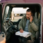 Owner operator in his truck cab on a broker call while filling out load paperwork, handling his own dispatcher work